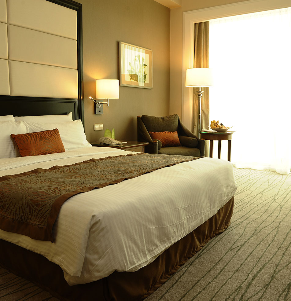 Hotel bedroom with a large bed with white and brown linens, warm lamps, armchair, and bright window light