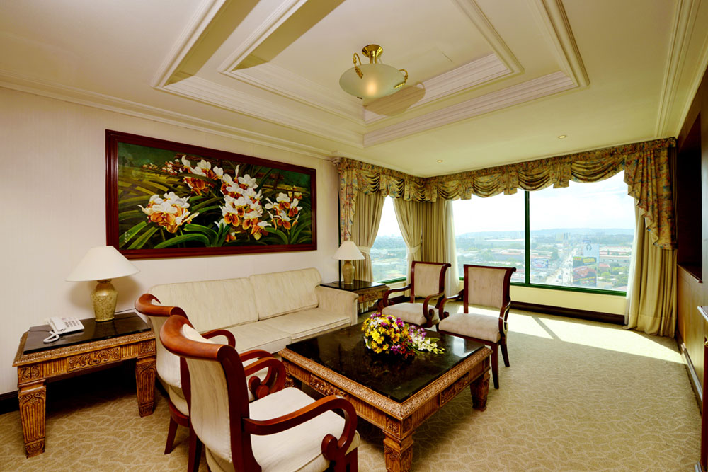 A hotel-style living room with a cream sofa, wooden furniture, a floral painting, and large windows showing a city view in warm tones