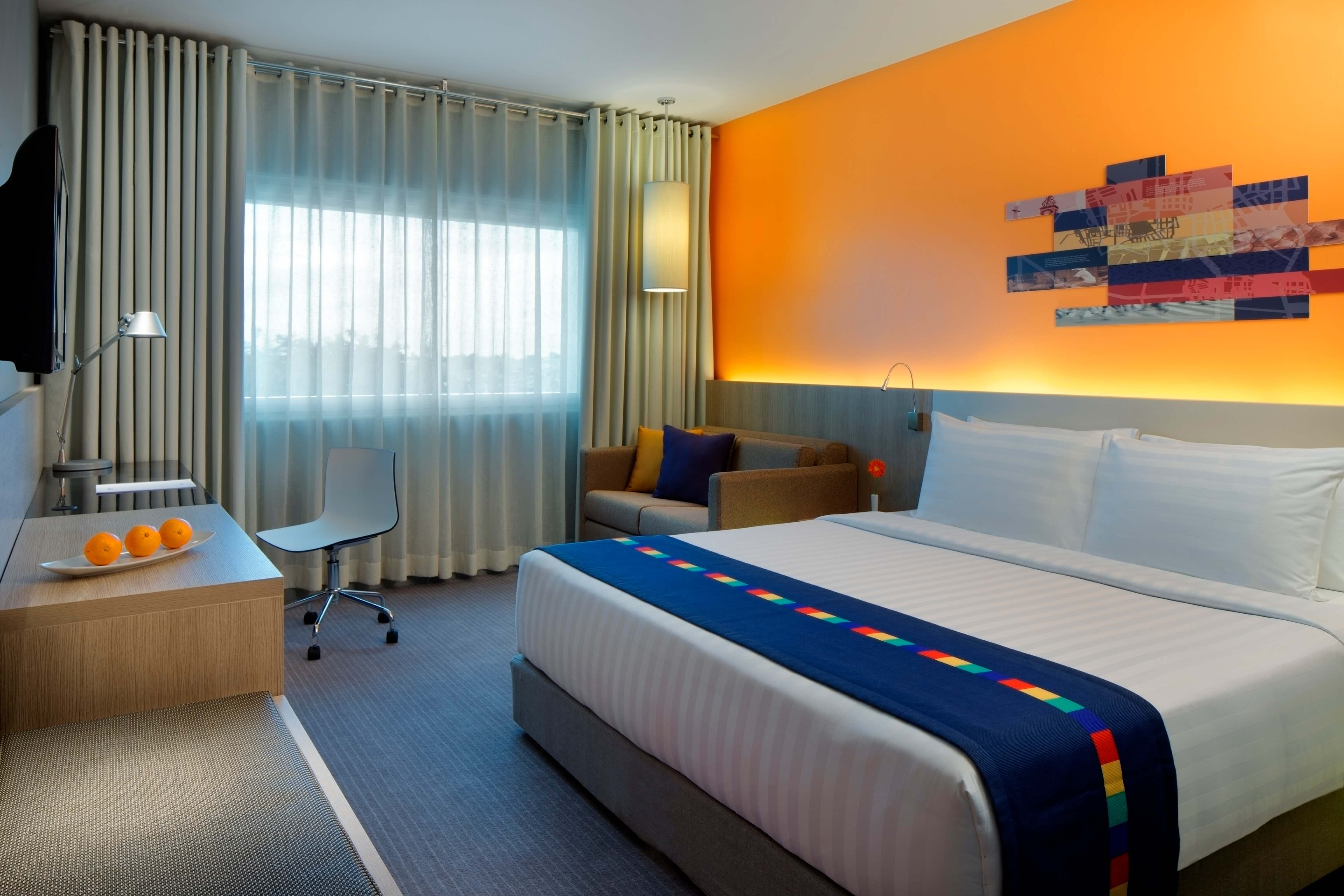 Modern hotel room with orange accent wall, white bed with colorful striped runner, and warm natural light